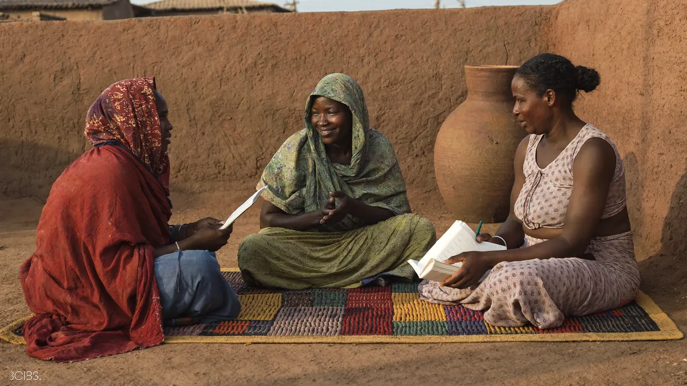 Trois femmes assises sur une natte, en conversation, l’une prenant des notes